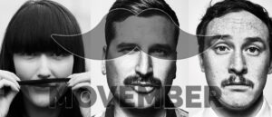 Two men and one woman standing side-by-side, each wearing a Movember moustache sticker and casual workplace attire, smiling and looking supportive in a bright modern office setting.