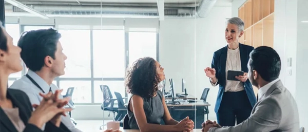 A professional female leader with short grey hair stands in a modern office meeting room, holding a digital tablet and presenting to a diverse, seated team of corporate professionals who are listening attentively.