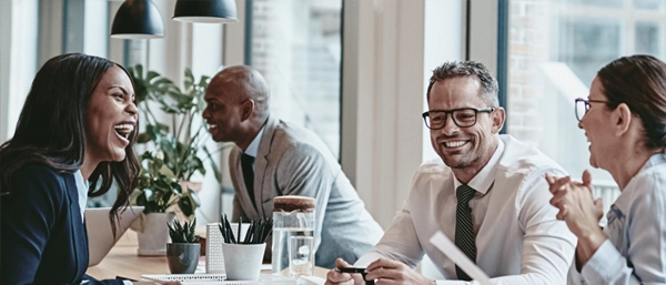 A group of diverse professionals laughing and collaborating around a meeting table in a bright, modern office. The team, including a woman laughing joyfully and a man in a white shirt and tie smiling, demonstrates a high-functioning work environment where psychosocial risks are successfully managed and controlled.
