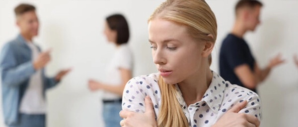 A concerned blonde woman wearing a polka-dot shirt stands in the foreground with her arms crossed tightly, looking downward in deep thought. In the softly blurred background, other small business employees are talking and gesturing in pairs, emphasizing a feeling of workplace stress, isolation, or overwhelming pressure.