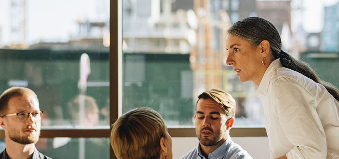 A professional team meeting in a conference room, with a confident female leader presenting to the group.