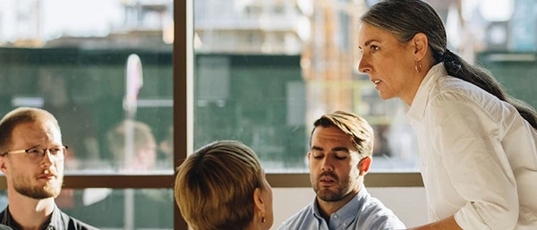 A professional woman with tied-back hair stands in profile, speaking calmly and authoritatively to three attentive corporate colleagues sitting before her in front of a large office window looking out onto a bright city background.