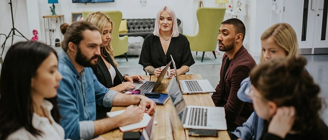 A diverse group of seven creative and corporate professionals gathered around a long wooden table in a stylish, modern workspace. Several open laptops are placed on the table, and team members are actively collaborating, writing in notebooks, and discussing strategies, with a woman with pink hair sitting at the centre of the group.