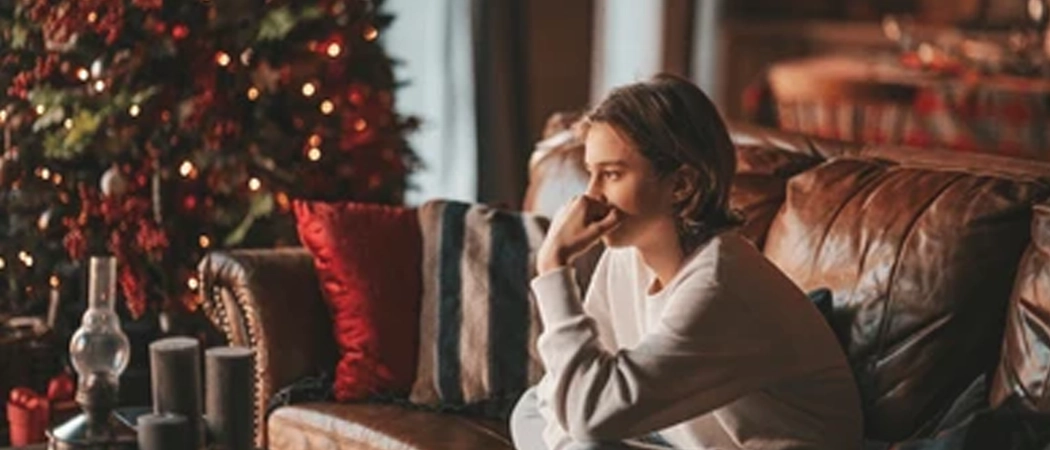 A person sits quietly on a brown leather couch, resting their chin on their hand in deep, pensive reflection. In the background, a beautifully lit Christmas tree glows softly next to a cozy, dimly lit dining room setup, capturing a quiet and contemplative moment during the holiday season.