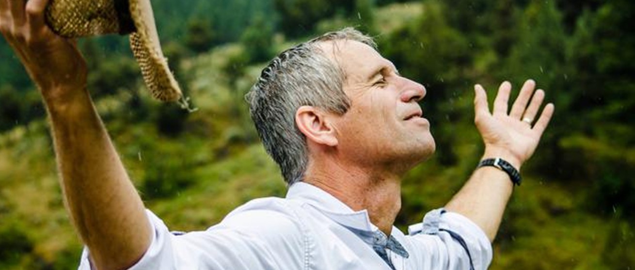 A side profile of a middle-aged man with grey hair, standing outdoors in a gentle rain shower with a lush green hillside in the background. His eyes are closed in peaceful reflection, his head is tilted up toward the sky, and his arms are spread wide open with one hand holding a straw hat, embodying freedom, thankfulness, and emotional release.