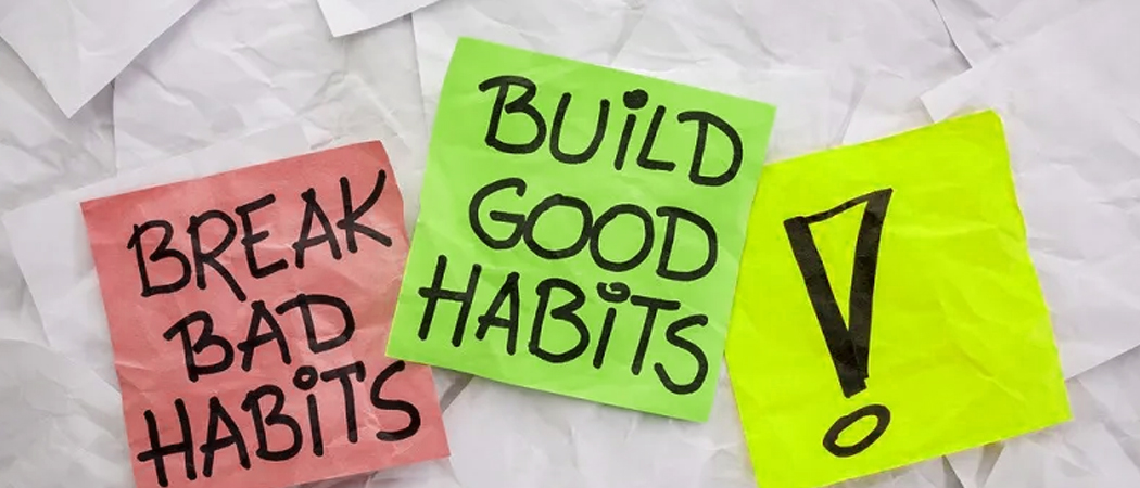 Three brightly colored sticky notes placed on a background of crumpled white paper. The left note is pink and reads "BREAK BAD HABITS," the center note is green and reads "BUILD GOOD HABITS," and the right note is neon yellow featuring a large black exclamation mark, symbolising personal development and behavioural change.