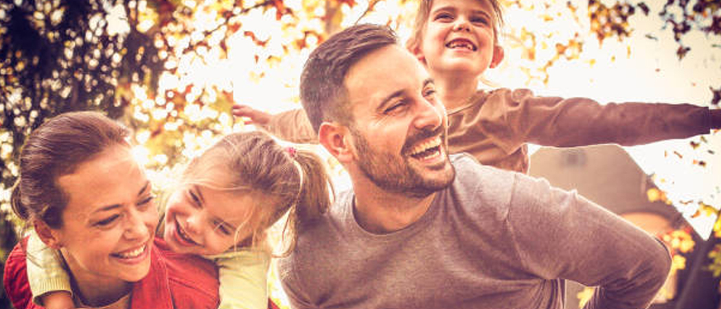 A joyful family enjoying an autumn day outdoors with golden leaves in the background. A smiling mother gives a piggyback ride to her young daughter, while beside them, a laughing father gives a piggyback ride to another cheering young child with outstretched arms, capturing a pure moment of happiness and connection.