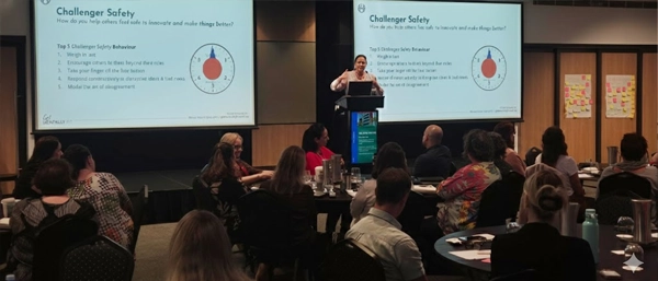 A professional speaker presenting a "Challenger Safety" workshop to a large audience. Two projector screens display "Top 5 Challenger Safety Behaviours," including "Weigh in early" and "Model the art of disagreement." The audience is seated at round tables, engaged in a training session about fostering psychological safety through everyday leadership actions.