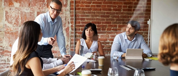 An older male manager with glasses stands at the head of a conference table in an office with a brick wall background, leaning forward and pointing to emphasise a point during a meeting with a focused group of seated male and female professionals.