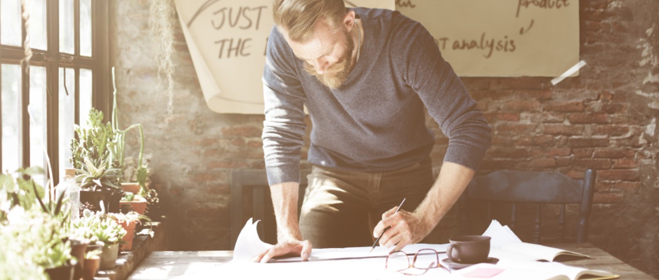 A professional man with a beard stands over a large wooden desk in a bright, rustic office space, leaning forward with deep concentration as he uses a pencil and a ruler to draw plans on a large sheet of paper. Potted green plants line the window to his left, creating a calm and intentional environment.