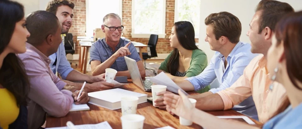 A diverse group of eight corporate professionals sitting around a long wooden table in a bright, modern loft office with brick accents. An older man wearing glasses and a blue shirt points and gestures enthusiastically while leading a vibrant team discussion, with laptops, coffee cups, and papers scattered across the table.