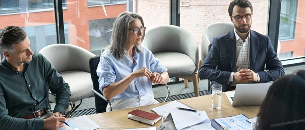 A senior female leader with grey hair and glasses leads a boardroom discussion with two male colleagues. They are seated at a wooden table with notebooks, a laptop, and data reports in a modern office with large glass windows, illustrating a serious executive consultation on workplace health and safety (WHS) strategy.