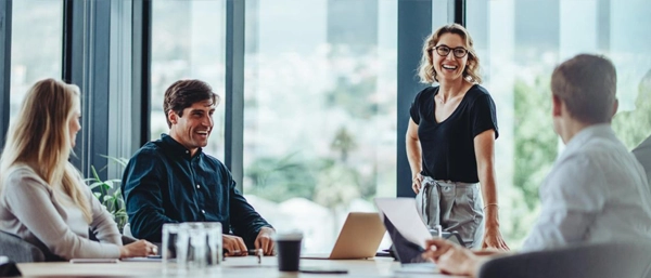 A professional woman with glasses and short blonde hair stands smiling and leading a team meeting in a bright, modern corporate office with floor-to-ceiling windows looking out over a cityscape. Three colleagues sit around the table, listening and smiling responsively.