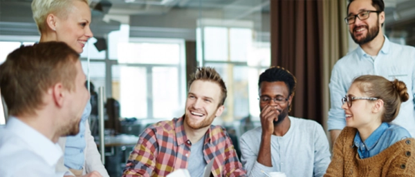 A diverse group of modern office professionals gathered around a table, laughing and smiling together during a positive team meeting in a bright, open-plan corporate workplace.
