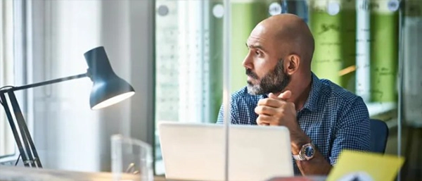 A professional, bald man with a neat beard sits at a modern office desk with his hands clasped, looking thoughtfully out a window. An open white laptop and a black desk lamp sit in front of him, separated by a glass partition that reflects a bright green wall in the background.