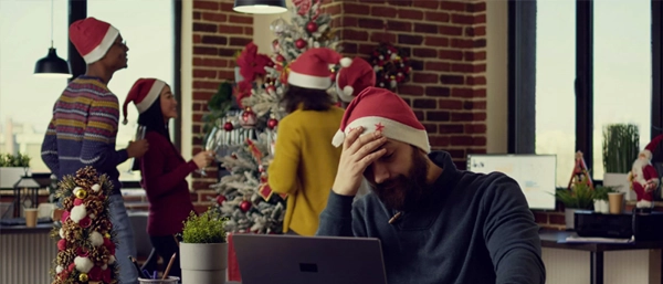 Stressed businessman in a Santa hat working at his laptop while colleagues celebrate the holiday season.