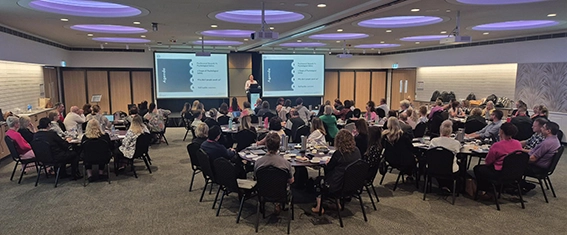 A large group of participants seated at round tables in a conference room during a workplace mental health and staff training presentation.