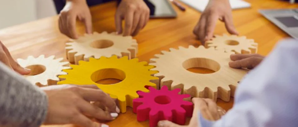 An overhead close-up shot of multiple hands working collaboratively around a wooden table to fit together large interlocking gears made of light natural wood, bright yellow, and vibrant pink.