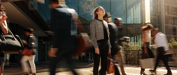 A professional woman in a grey checkered blazer stands still and looks upward with a thoughtful expression in a busy, sunlit city plaza. Around her, several commuters are captured as blurred figures in motion, creating a visual contrast between her moment of strategic reflection and the fast-paced urban environment.