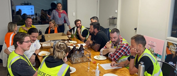 A group of workers in high-visibility vests sitting around a wooden table in a casual meeting room during a meal break. A woman stands at the head of the table gesturing as she speaks to the group, illustrating an informal but critical leadership moment regarding safety and risk management in a frontline or industrial setting.